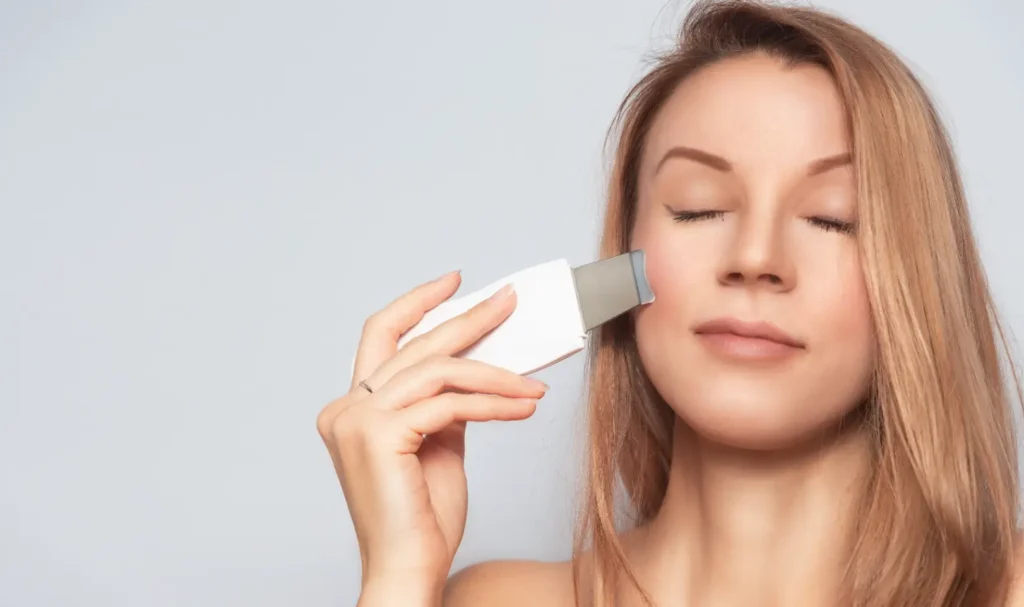 Woman holding a handheld skincare device against her cheek with eyes closed.