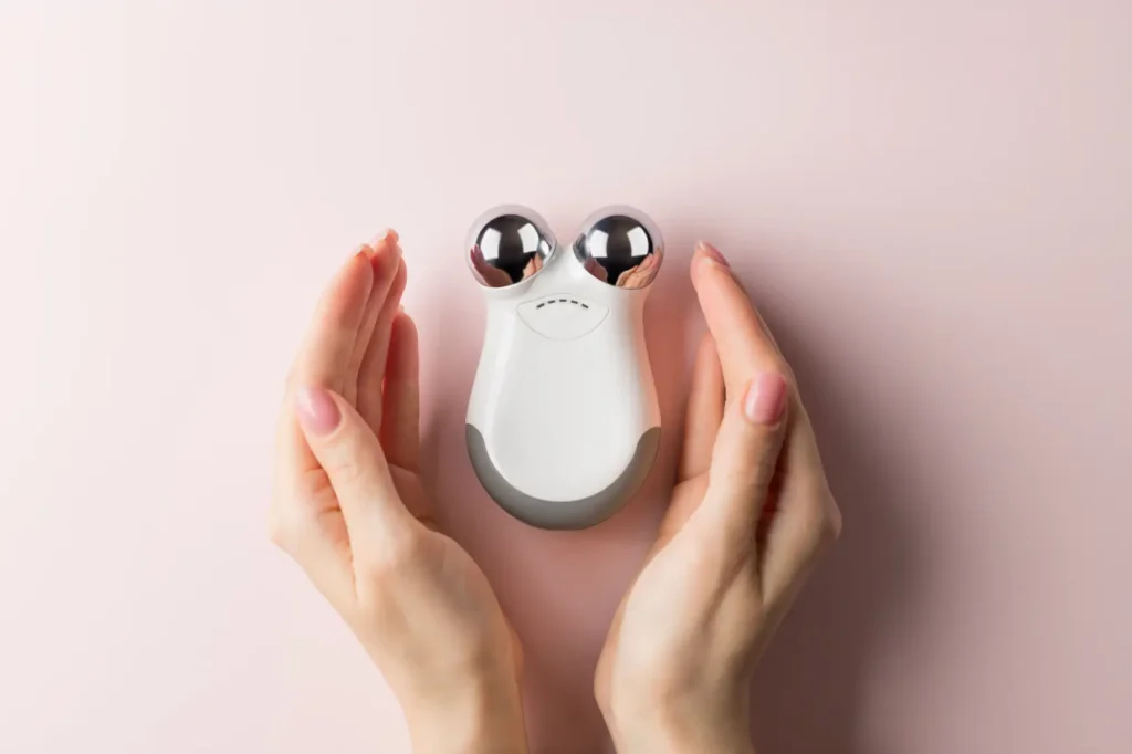 Hands holding a dual-roller facial massage device on a pink background.