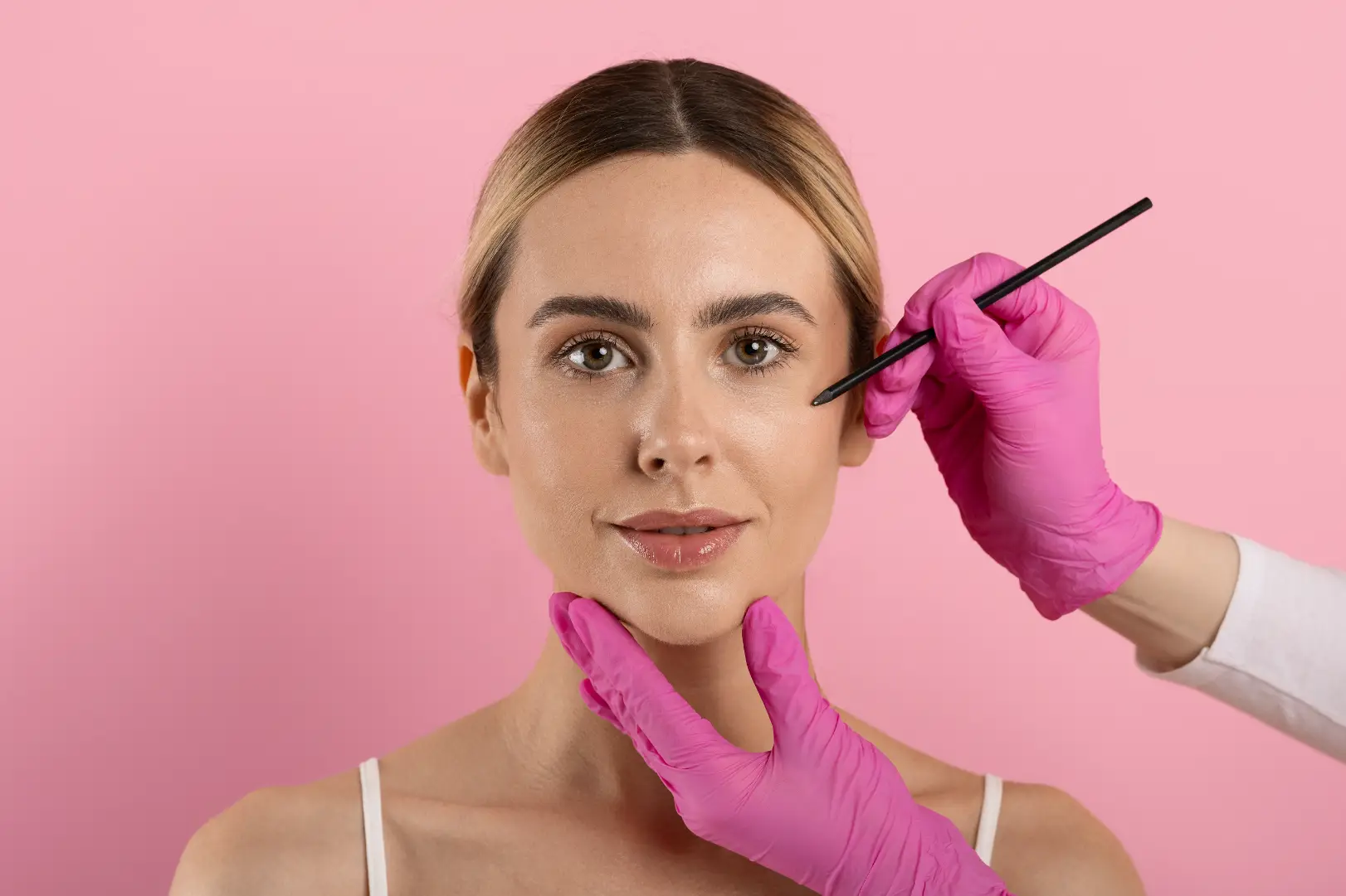 Woman faces forward while gloved hands hold her chin and point a black tool toward her cheek.