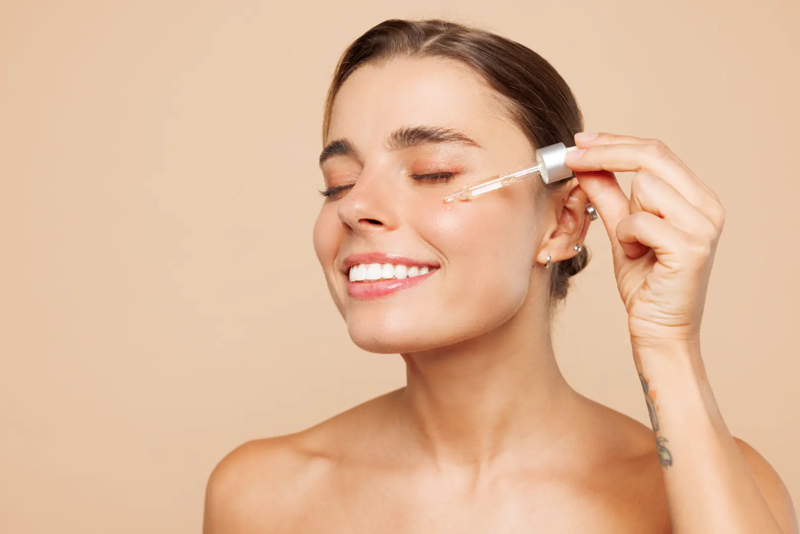 Woman applies clear serum to her cheek with a glass dropper against a beige background.