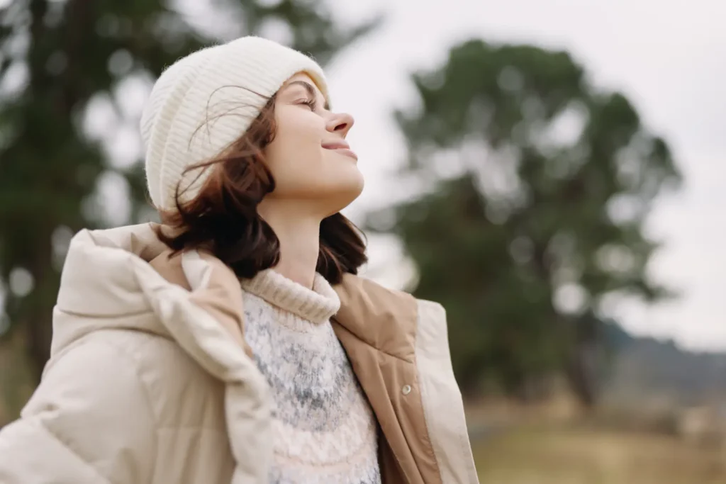 Woman in a knit hat and beige coat stands outdoors with her eyes closed and trees in the background.