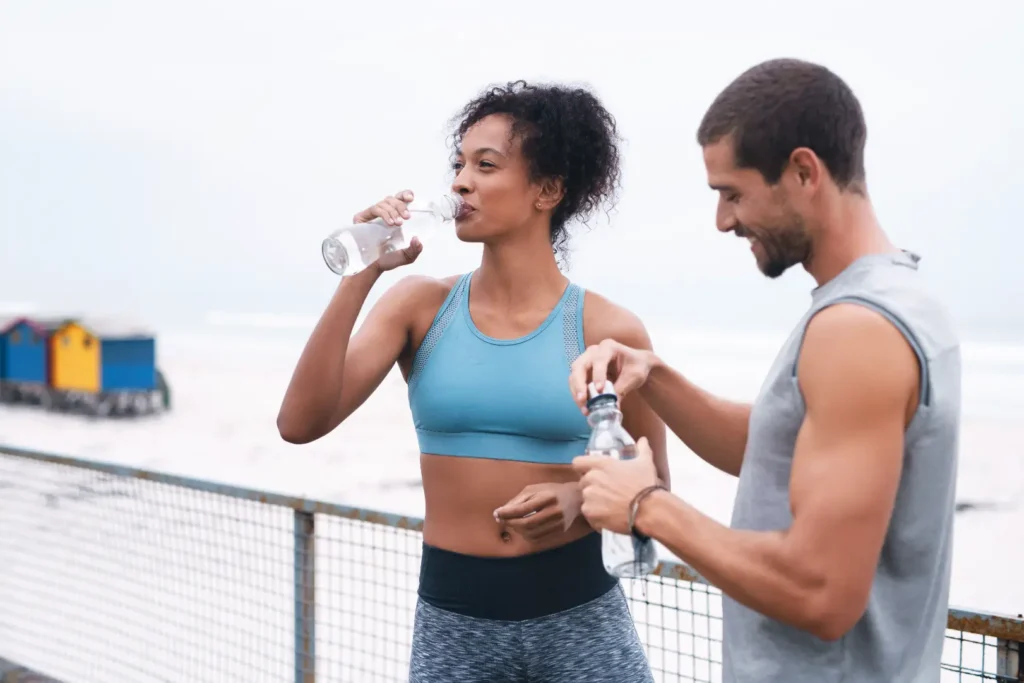 Woman in athletic wear drinks bottled water while a man beside her opens another bottle outdoors.