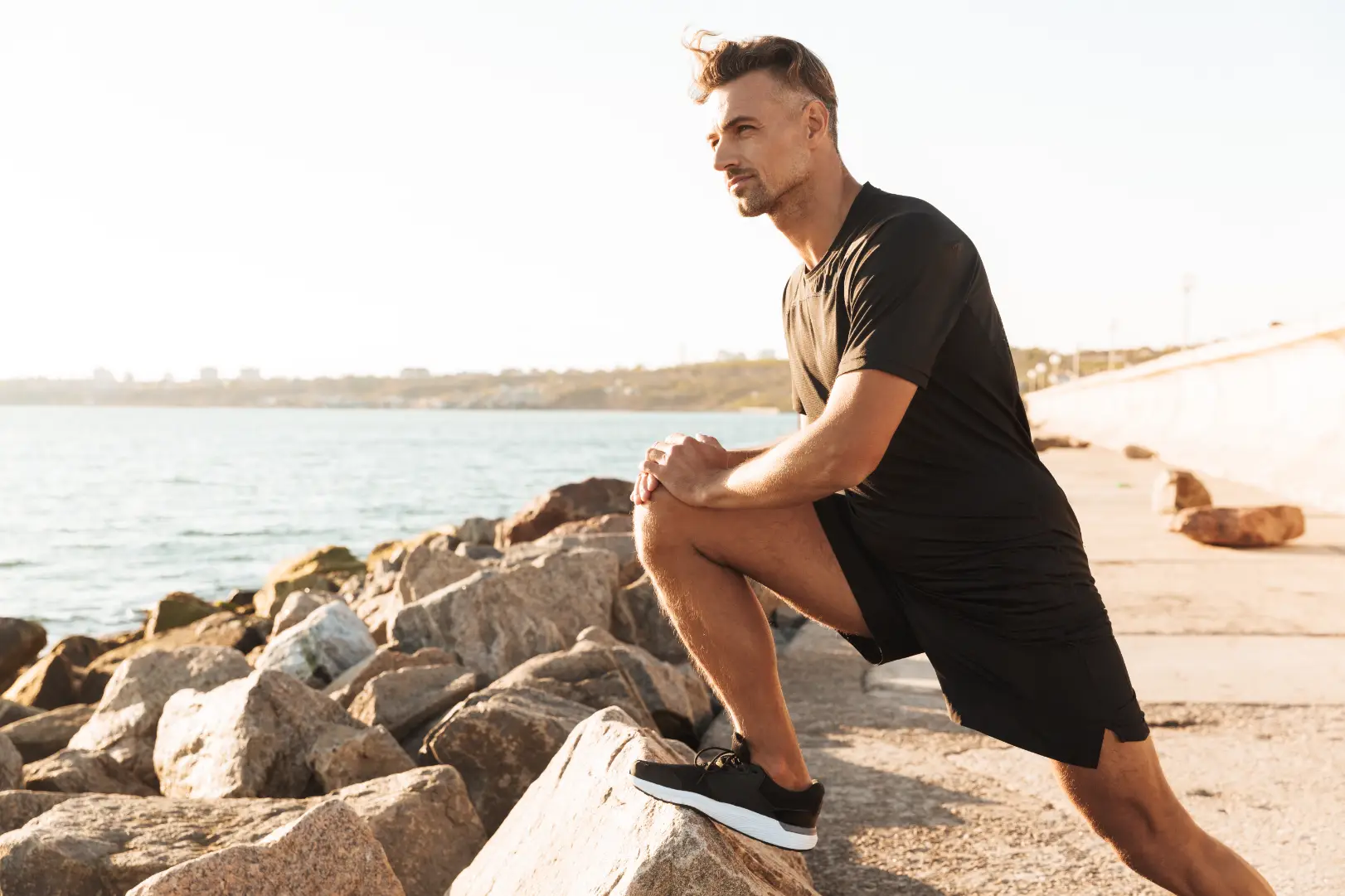 Man in black athletic clothing stretches one leg on a rock beside the water in warm sunlight.