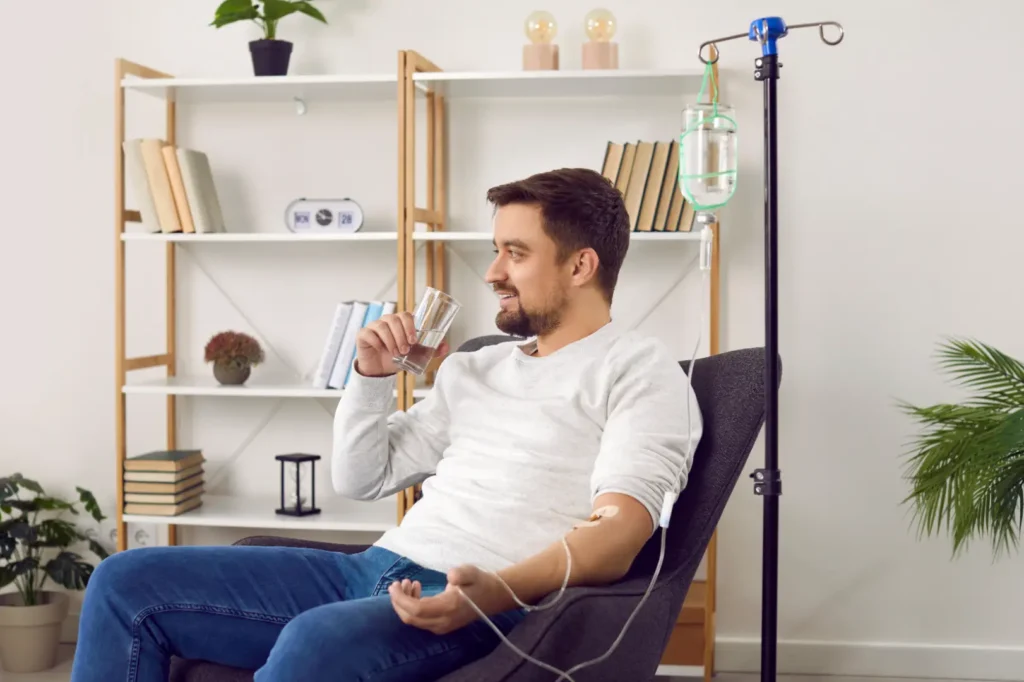Man sits in a chair drinking water with an IV line connected to his arm and an infusion bag beside him.