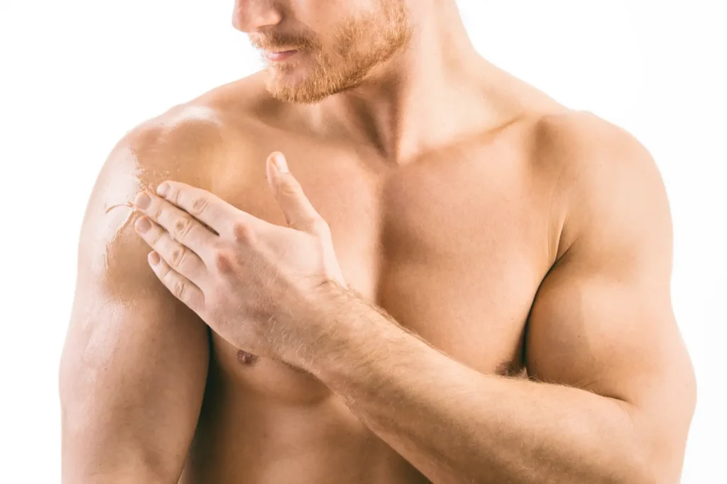 Shirtless man applies a clear topical product to his shoulder against a plain white background.