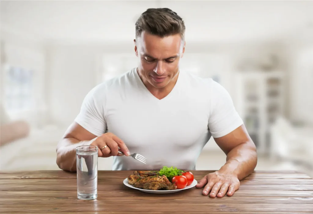 Muscular man in a white shirt sits at a table with grilled chicken, lettuce, tomatoes, and a glass of water.