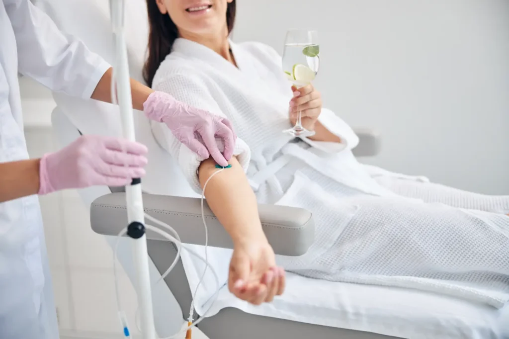 Woman in a white robe holds a glass of water while a clinician adjusts an IV line attached to her arm.